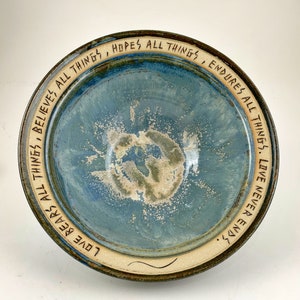 May include: A blue ceramic bowl with a white rim and a brown glaze. The bowl has a quote inscribed around the rim: "Love bears all things, believes all things, hopes all things, endures all things. Love never ends."
