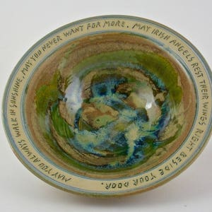 May include: A ceramic bowl with a blue, green, and brown glaze. The bowl has a white rim with an inscription that reads: "MAY IRISH ANGELS REST THEIR WINGS RIGHT BESIDE YOUR DOOR. ALWAYS WALK IN SUNSHINE, MAY YOU NEVER WANT FOR MORE. MAY YOU ALW"