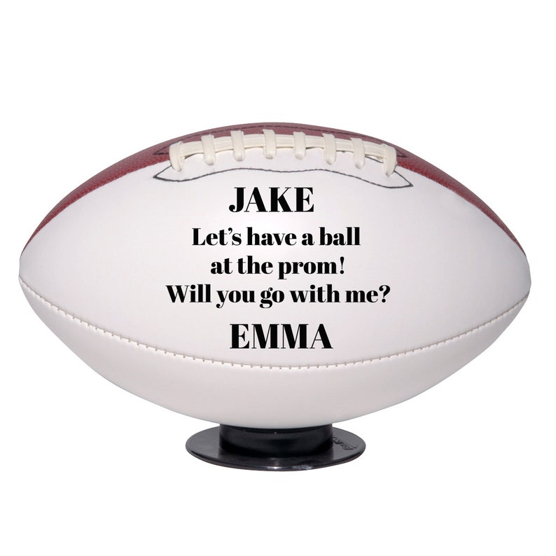 May include: White football with black text that reads "JAKE Let's have a ball at the prom! Will you go with me? EMMA".