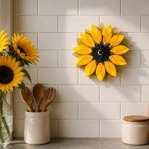 Op de afbeelding: Een zonnebloemvormige wandklok met heldergele bloemblaadjes en een donker centrum, voorzien van zwarte wijzers. De klok hangt aan een witte tegelmuur naast een glazen vaas gevuld met verse zonnebloemen, houten keukengerei en diverse keukenartikelen.