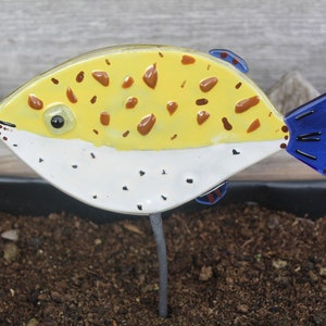 Puede incluir: Una estaca de jardín de pez globo de vidrio amarillo, blanco y azul. El pez tiene manchas marrones y una aleta caudal azul.