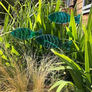 May include: Three teal blue glass fish sculptures with white lines are placed in a garden setting. The fish are on metal stakes and are surrounded by green grass and foliage.