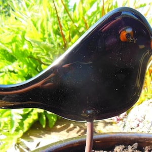 以下が含まれることがあります： オレンジ色のくちばしと目を持つ、光沢のある黒いガラスの鳥の彫刻。金属製の棒に取り付けられています。鳥は土の入った鉢に止まっており、背景はぼやけた緑色です。