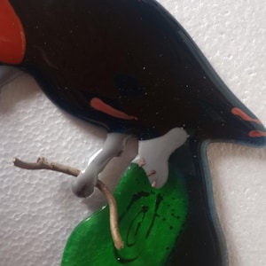 May include: A decorative bird figurine with a glossy black body, red accents, and grey legs. It is perched on a green leaf with a whimsical face and a small brown twig. The bird is a unique piece of art.