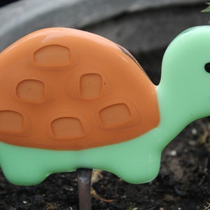 Puede incluir: Una figurita de tortuga de vidrio verde y naranja con una cara sonriente y una concha detallada. La tortuga está sentada sobre una superficie marrón.
