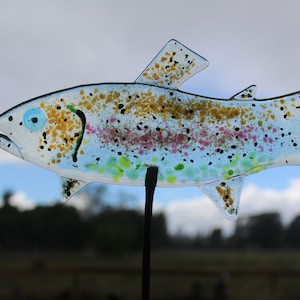 Puede incluir: Una escultura de pez de vidrio con un cuerpo transparente y manchas marrones, rosadas, verdes y negras. El pez está montado en una varilla de metal negro.