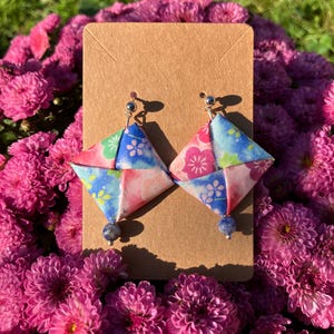 May include: A pair of origami earrings with a blue, pink, and green floral pattern. The earrings are made of paper and have a blue gemstone dangle.