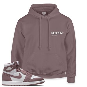 May include: A pair of white and dusty rose sneakers with a white swoosh logo and a white Jordan logo. The sneakers are paired with a dusty rose hoodie with the text "REDRUM" and "REDRUM NATION" printed in white.