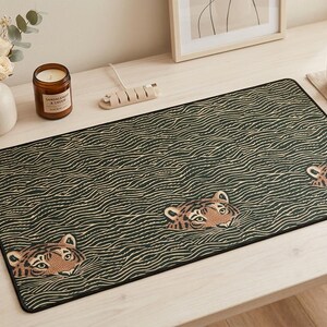 May include: A rectangular desk mat with a black border and a dark green wave pattern. The mat features three tiger faces. The desk mat is on a light-coloured wooden desk, with a candle, flowers, and a cup of coffee.