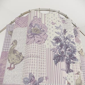 May include: A patchwork shower curtain with a repeating pattern of ducks, flowers, and botanical illustrations in lavender, grey, and white. The curtain is hung on a curved metal rod.