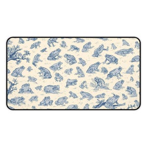 Pode incluir: Um mousepad retangular com um padrão repetitivo de ilustrações de sapos azuis em um fundo creme. Os sapos variam em tamanho e pose, criando um design caprichoso. Um galho com folhas está no canto superior esquerdo.