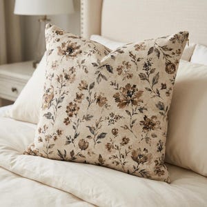 May include: A square scatter cushion featuring a cream-coloured base with a repeating botanical print of flowers and leaves in shades of brown, tan, and muted grey. The cushion has a visible zip closure along the side edge and a soft, textured fabric.