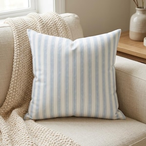 May include: Square throw pillow with a light blue and white striped pattern. The pillow is on a beige sofa with a cream-colored knitted throw blanket. The pillow has a classic, farmhouse style.