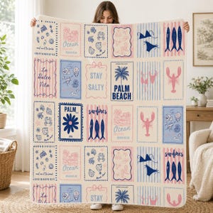 May include: A cream-coloured throw blanket featuring a grid of rectangular panels with blue and pink illustrations of palm trees, lobsters, sardines, seashells, bikinis, and text including Palm Beach, Stay Salty, and La Dolce Vita.