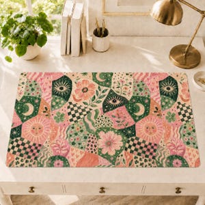 May include: A rectangular desk mat featuring a repeating pattern of pink flowers, green foliage, suns with faces, crescent moons, eyes, and black and white checkered sections. The design incorporates various organic shapes and small heart motifs.