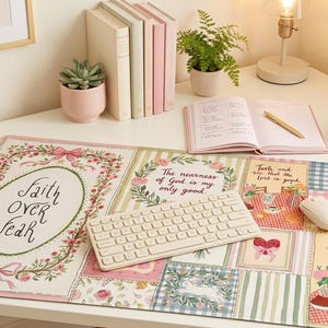 May include: A decorative desk mat with a floral and patchwork design. The mat features the text "Faith Over Fear" and other inspirational quotes. A wireless keyboard and mouse sit on the mat, alongside a notebook, pen, and a cup and saucer.