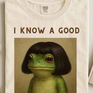 May include: Cream-colored t-shirt featuring the phrase "I KNOW A GOOD" above a whimsical illustration of a green frog with dark brown hair. The shirt has a classic crew neck and a tag.