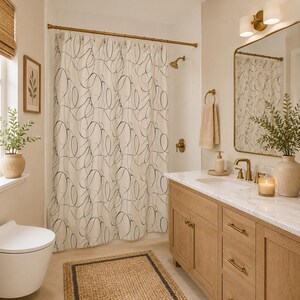 May include: A shower curtain featuring a continuous black line drawing pattern on a cream background, suspended from a brass rod. The curtain hangs in a bathroom with a light wood vanity, a marble countertop, and a matching brass faucet fixture.