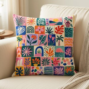 May include: A square throw pillow featuring a grid of colorful painted squares. Each square displays distinct botanical, floral, or landscape motifs in vibrant shades of pink, orange, blue, green, and yellow against a cream-colored background.