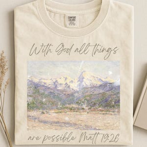 May include: A cream-coloured t-shirt featuring a landscape painting of mountains and the text "With God all things are possible Matt 19:26." The shirt has a crew neck and short sleeves. The shirt has a tag that says "Comfort Colors."