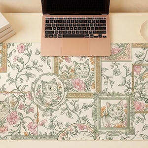 Op de afbeelding: Een crèmekleurige bureaumat met ingelijste kattenportretten, bloempatronen en een illustratie van een slapende kat. Een laptop, boeken, een pen, een kopje thee en een vaas met bloemen staan op het bureau. De mat heeft roze en groene accenten.