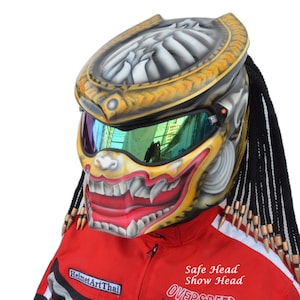 May include: A gold and black motorcycle helmet with a predator-like design and a red mouth. The helmet has a green visor and black dreadlocks.