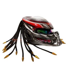 Pu&ograve; includere: Un casco da moto personalizzato con un design Predator. Il casco presenta una combinazione di colori argento e rosso, con dreadlocks neri e punte color oro. La visiera &egrave; colorata e il casco ha un aspetto dettagliato e strutturato.