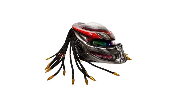 Pu&ograve; includere: Un casco da moto personalizzato con un design Predator. Il casco presenta una combinazione di colori argento e rosso, con dreadlocks neri e punte color oro. La visiera &egrave; colorata e il casco ha un aspetto dettagliato e strutturato.