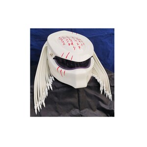 May include: A white motorcycle helmet with a design inspired by the Predator. The helmet features a dark visor, red markings, and white dreadlocks with pointed tips. The top of the helmet has red symbols.