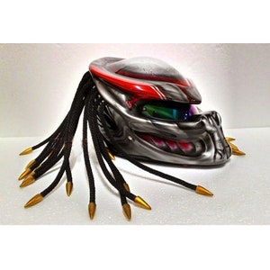 Pu&ograve; includere: Un casco da moto personalizzato con un design Predator. Il casco presenta una combinazione di colori grigio metallizzato e rosso, con dreadlocks neri e punte dorate. La visiera ha una tinta verde e blu.