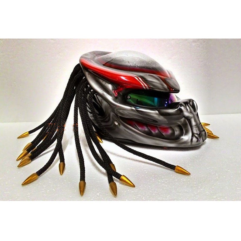 Pu&ograve; includere: Un casco da moto personalizzato con un design Predator. Il casco presenta una combinazione di colori grigio metallizzato e rosso, con dreadlocks neri e punte dorate. La visiera ha una tinta verde e blu.