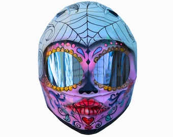 May include: A motorcycle helmet with a Day of the Dead sugar skull design. The helmet features a white top with a spiderweb design, a pink and purple face with gold-rimmed eye sockets, and red lips. The visor reflects the surroundings.