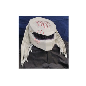 May include: A white motorcycle helmet with a design inspired by the Predator movie franchise. The helmet features a dark visor, red markings, and long, white dreadlocks. The helmet is designed for motorcycle use.