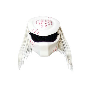 May include: White motorcycle helmet with a Predator design. The helmet features a smooth, glossy finish, dark visor, and red accents. It has white dreadlocks and a unique, angular shape, perfect for cosplay or themed events.