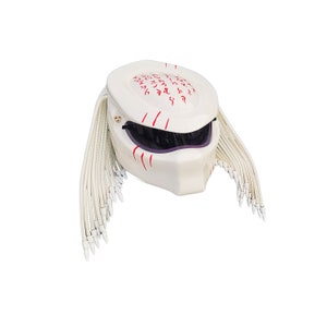 May include: A white motorcycle helmet with a Predator design. The helmet features a dark visor, red markings, and white dreadlocks. The top of the helmet has red markings and a gold emblem.