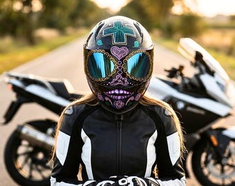 May include: A black motorcycle helmet featuring a colorful sugar skull design with a teal cross on the forehead and reflective blue visors. The helmet is worn with a black and white leather motorcycle jacket, positioned in front of a white motorcycle.