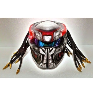 Pu&ograve; includere: Un casco da moto personalizzato con un design Predator. Il casco presenta una combinazione di colori rosso e argento con dreadlocks neri e punte dorate. La visiera ha una finitura a specchio multicolore.