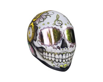 May include: A white motorcycle helmet with a skull design. The helmet features a detailed sugar skull design with floral and musical note accents. The visor has a reflective gold tint. The helmet is a full-face style.