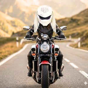 May include: A person riding a red and silver motorcycle on a paved road. The person is wearing a white helmet with long, white, fringed details and a black leather jacket. The motorcycle has a large headlight and is moving forward.