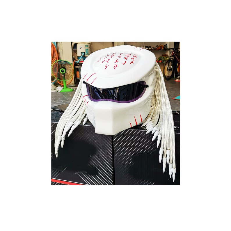 May include: White motorcycle helmet with a Predator design. The helmet features a visor, red markings, and long white dreadlocks. The helmet is sitting on a black and silver patterned surface.