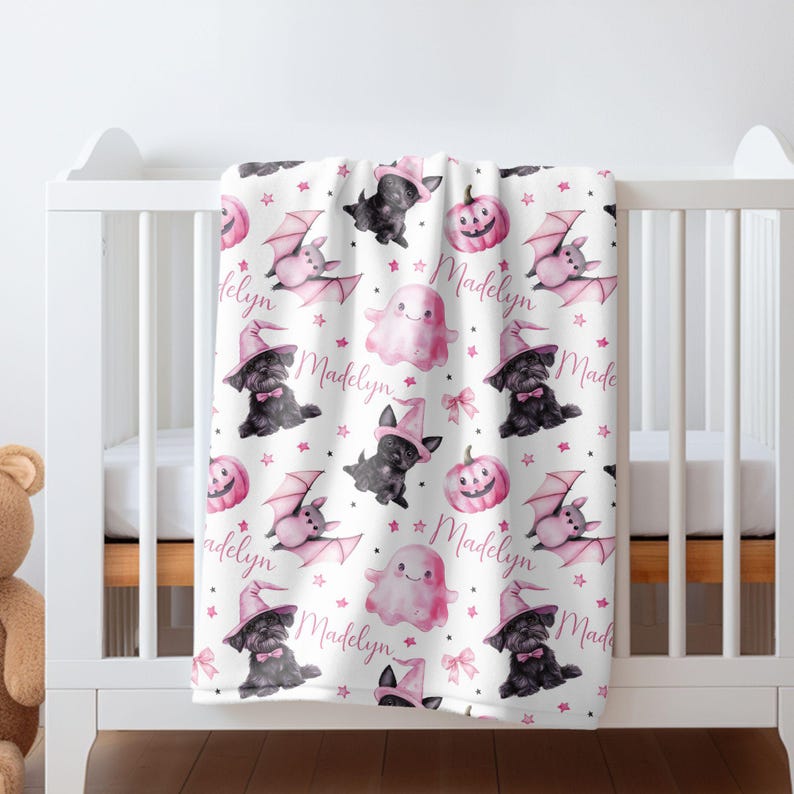 May include: A pink and black Halloween themed baby blanket with a repeating pattern of bats, ghosts, pumpkins, and witches. The blanket features the name "Madelyn" repeated throughout the design.