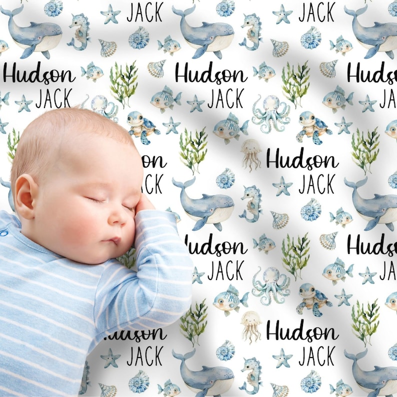 May include: A white baby blanket with a blue and green watercolor pattern featuring whales, seahorses, fish, and seashells. The text "Hudson Jack" is repeated throughout the blanket.