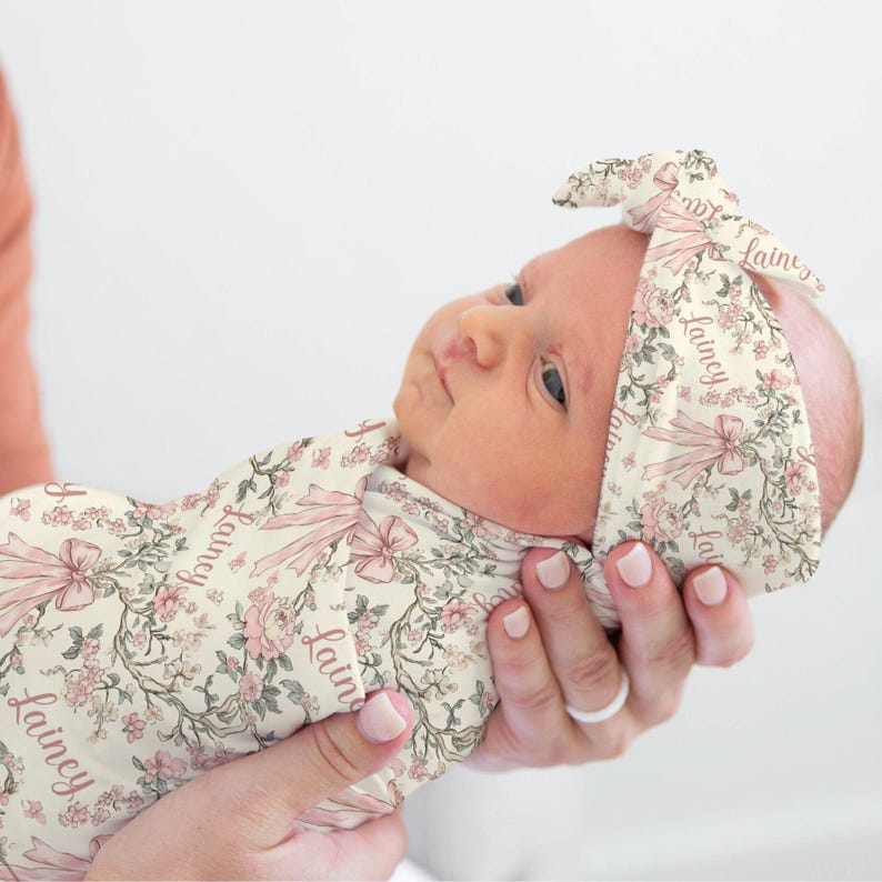 May include: A newborn baby wrapped in a floral swaddle and matching headband. The cream-colored fabric features pink flowers, bows, and the name "Lainey" in script. The baby is held in someone's arms.
