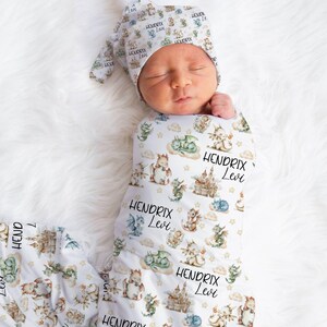 May include: A white swaddle blanket featuring a dragon and castle design. The blanket is decorated with the names "Hendrix Levi" repeated throughout the pattern. A baby is swaddled in the blanket and wearing a matching hat.