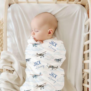 May include: A white swaddle blanket with a blue airplane pattern and the name "Jack Henry" repeated on the blanket. The blanket is wrapped around a baby sleeping in a crib.