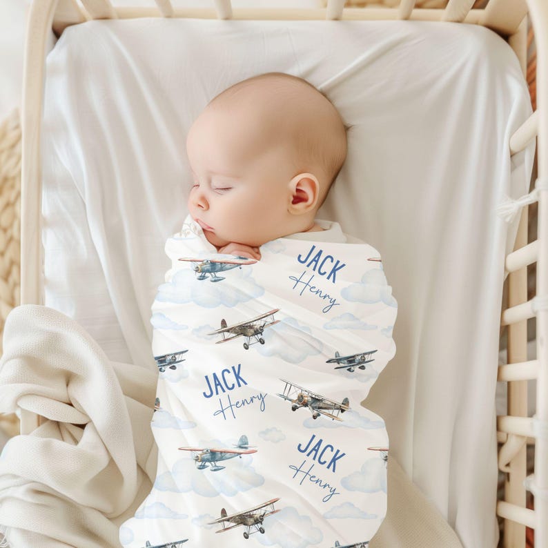 May include: A white swaddle blanket with a blue airplane pattern and the name "Jack Henry" repeated on the blanket. The blanket is wrapped around a baby sleeping in a crib.