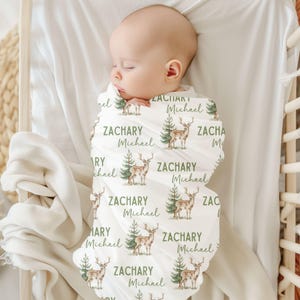 May include: A white swaddle blanket with a repeating pattern of green trees and brown deer. The blanket is personalized with the name "Zachary Michael" repeated throughout the design.