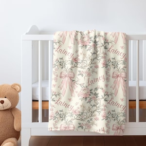 May include: A cream-colored baby blanket with a floral and bow pattern. The design features pink roses, bows, and the name "Lainey" in a cursive font. The blanket is draped over a white crib.