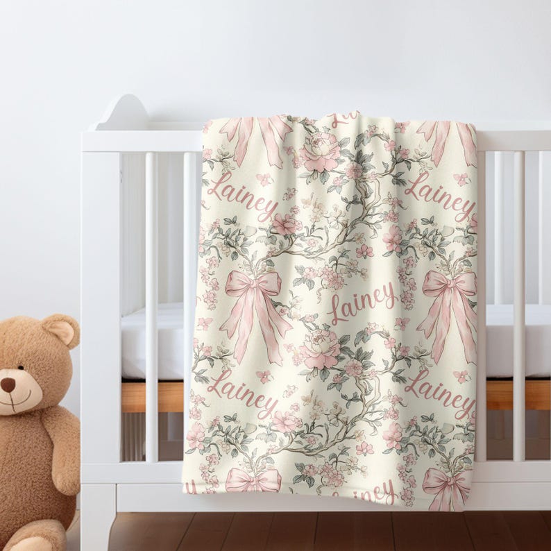 May include: A cream-colored baby blanket with a floral and bow pattern. The design features pink roses, bows, and the name "Lainey" in a cursive font. The blanket is draped over a white crib.