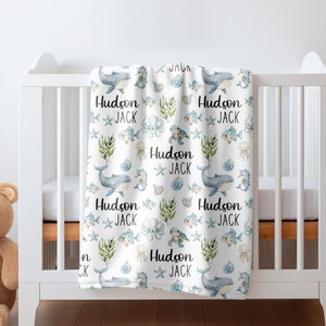 May include: A white baby blanket with a blue and green ocean-themed pattern. The blanket features whales, fish, seahorses, and seaweed. The text "Hudson Jack" is repeated throughout the pattern.